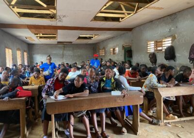 Students seated at wooden desks in a crowded classroom with a damaged ceiling while UNIMAC staff stand at the front