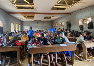 UNIMAC staff addressing a packed classroom of students seated at wooden benches under a damaged ceiling