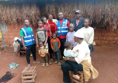 UNIMAC field officers standing with a family of adults and young children outside a home with a thatched wall