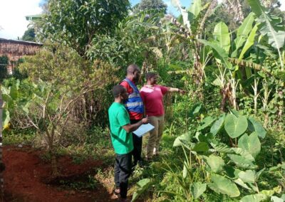 Two UNIMAC field officers in reflective vests reviewing a home garden with a community member who points out her crops