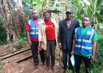 Two UNIMAC field officers in reflective vests standing with a woman and an elderly man in a banana farm