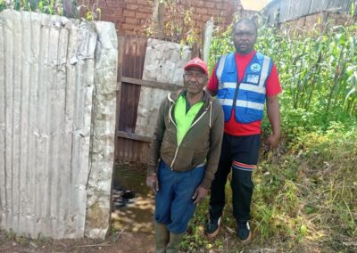 A UNIMAC field officer in a reflective vest standing with an elderly man in a red cap beside a household latrine