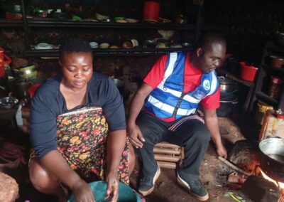 A UNIMAC field officer in a reflective vest seated beside a woman preparing food in her kitchen