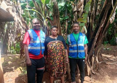 Two UNIMAC field officers in reflective vests standing with a woman in her banana plantation