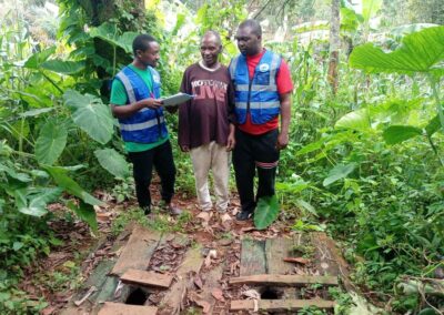 Two UNIMAC field officers and an elderly man standing beside an open pit latrine covered with wooden planks