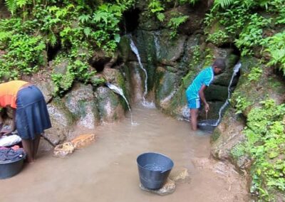 Two people collecting water at an open spring at the foot of a green hillside, with buckets in muddy water