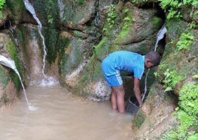 A young person bending to fill a bucket at an open spring flowing from a rocky hillside